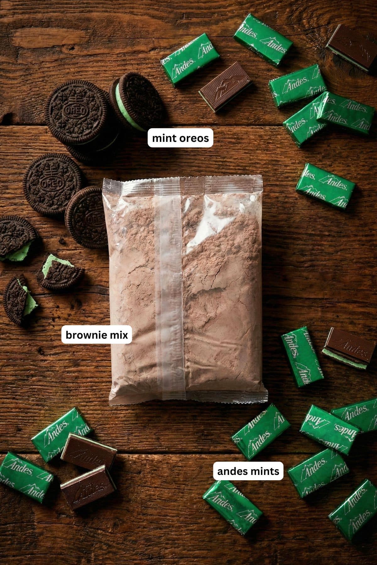 Fudgy chocolate brownies are filled with layers of Mint Oreos and Andes Mints to create these epic Andes Mint Brownies. Labeled ingredients for mint brownie recipe: a bag of brownie mix, mint Oreos, and individually wrapped Andes mints are spread out on a wooden surface.