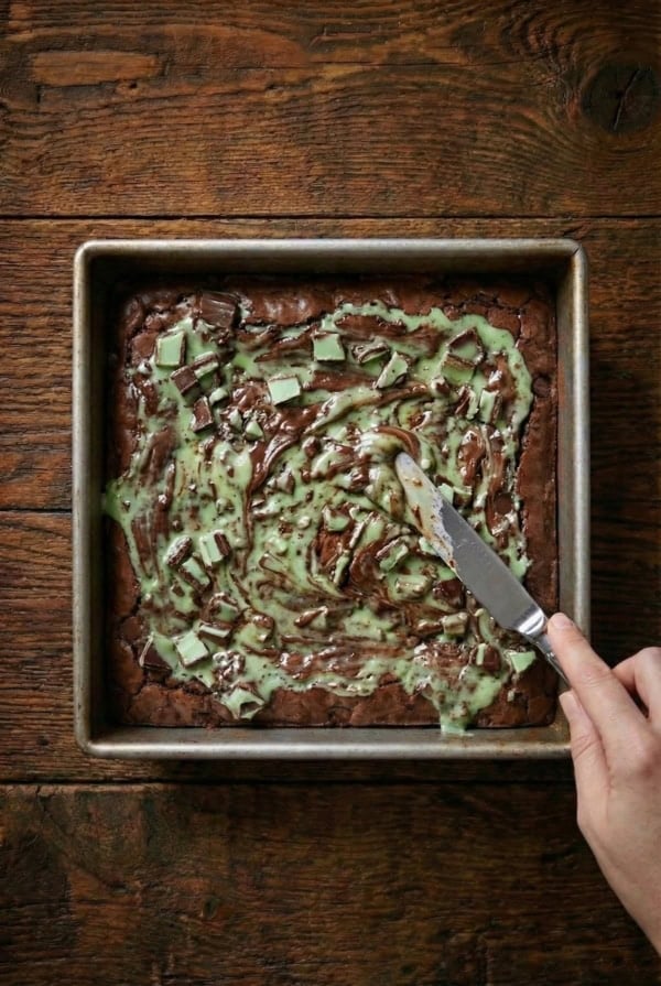 Fudgy chocolate brownies are filled with layers of Mint Oreos and Andes Mints to create these epic Andes Mint Brownies. A hand uses a knife to swirl melted andes mints on top of brownies in a square metal baking pan.
