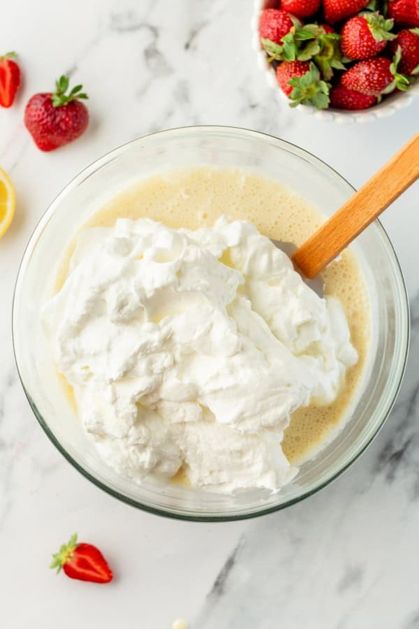 With no ice cream maker needed, this velvety Strawberry Cheesecake Ice Cream recipe has thick swirls of strawberry compote and graham cracker crumbs. A glass bowl filled with whipped cream being folded into a sweetened condensed milk mixture with a wooden spatula.