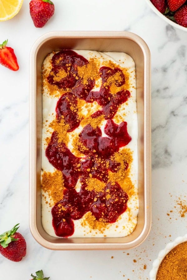 With no ice cream maker needed, this velvety Strawberry Cheesecake Ice Cream recipe has thick swirls of strawberry compote and graham cracker crumbs. A rectangular pan filled with a creamy no-churn ice cream mixture and topped with red strawberry compote and sprinkled with crushed graham crackers.