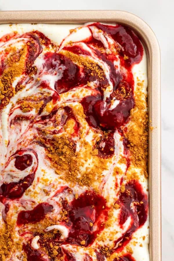 With no ice cream maker needed, this velvety Strawberry Cheesecake Ice Cream recipe has thick swirls of strawberry compote and graham cracker crumbs. A close-up of a rectangular dish filled with Strawberry Cheesecake Ice Cream, swirled with red strawberry sauce and sprinkled with crumbled graham crackers on top.