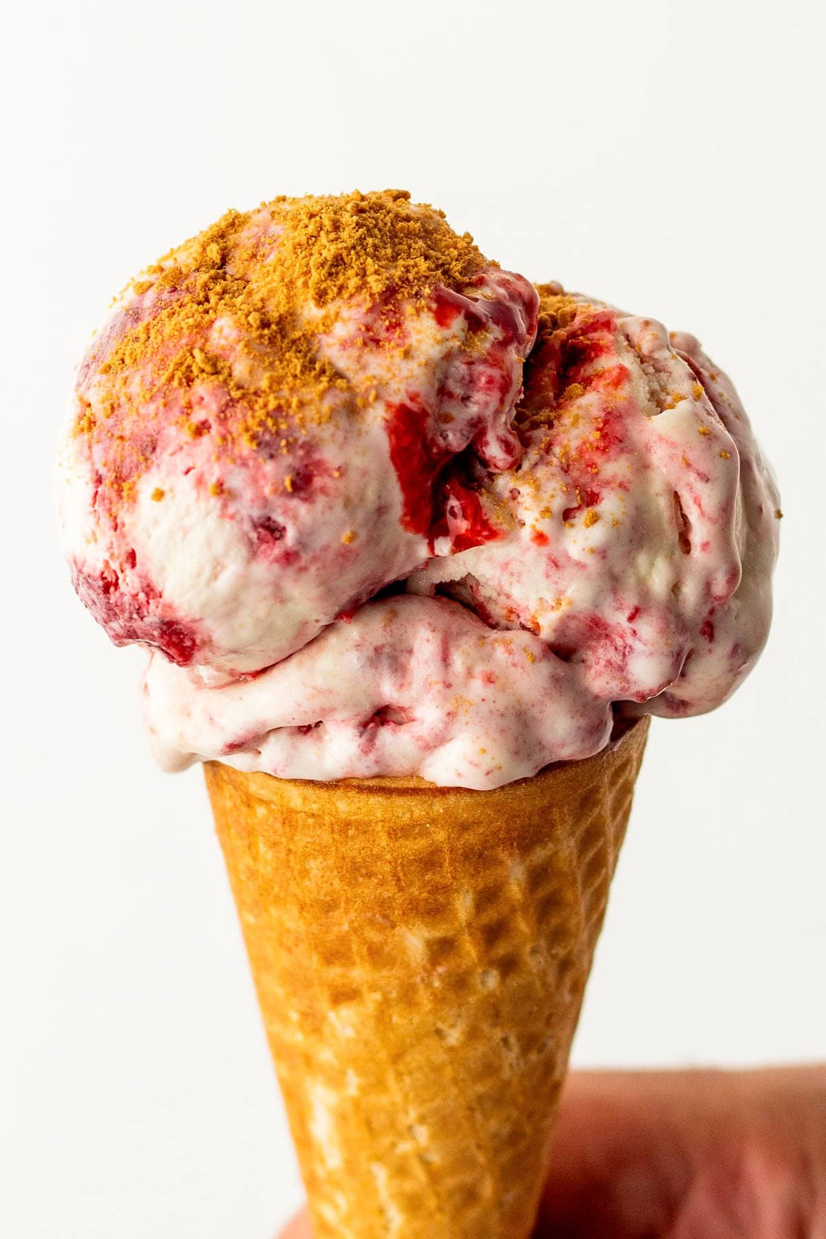 With no ice cream maker needed, this velvety Strawberry Cheesecake Ice Cream recipe has thick swirls of strawberry compote and graham cracker crumbs. A hand holds an ice cream cone filled with creamy, swirled strawberry cheesecake ice cream topped with a sprinkle of crumbled graham crackers.