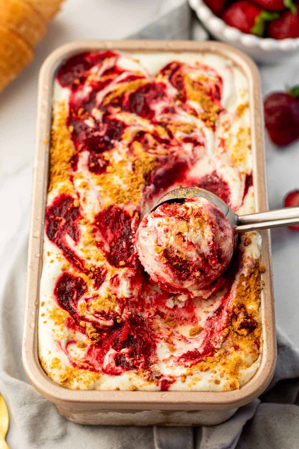 With no ice cream maker needed, this velvety Strawberry Cheesecake Ice Cream recipe has thick swirls of strawberry compote and graham cracker crumbs. A rectangular container of swirled Strawberry Cheesecake Ice Cream with a scoop taken out, showing creamy white ice cream with red strawberry swirls and crumbled graham cracker topping. A metal ice cream scoop rests in the container.