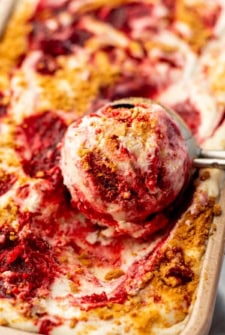 A metal ice cream scoop lifts a serving of creamy No-Churn Strawberry Cheesecake Ice Cream from a pan filled with more ice cream.