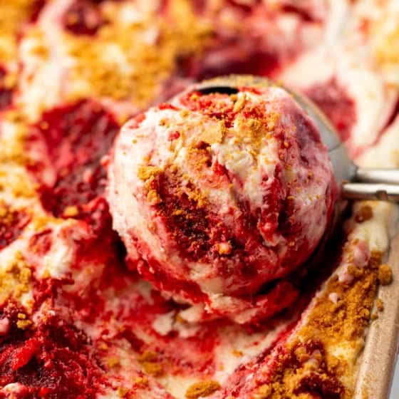 A metal ice cream scoop lifts a serving of creamy No-Churn Strawberry Cheesecake Ice Cream from a pan filled with more ice cream.