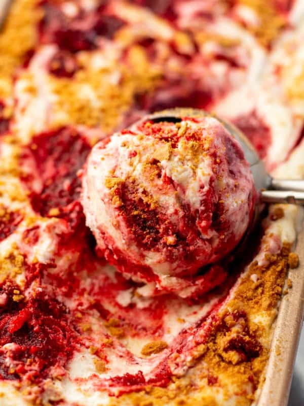 A metal ice cream scoop lifts a serving of creamy No-Churn Strawberry Cheesecake Ice Cream from a pan filled with more ice cream.