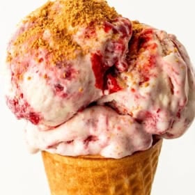A close-up of a waffle cone filled with strawberry cheesecake no-churn ice cream, topped with a crumbly graham cracker crust.