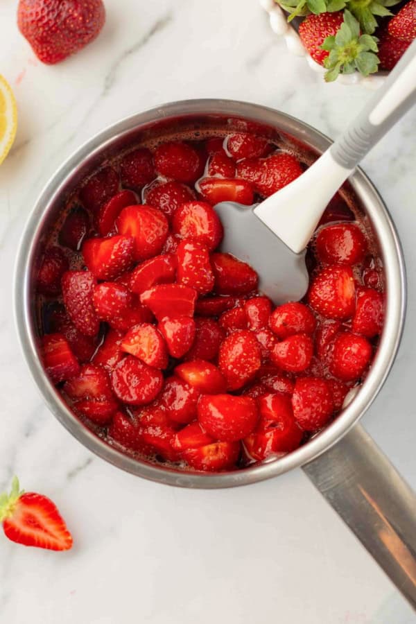 Made with only 4 ingredients in just 10 minutes, this homemade Strawberry Compote recipe is so thick and jammy - with no cornstarch needed! A saucepan filled with halved strawberries cooking in syrup being stirred with a gray-and-white spatula.