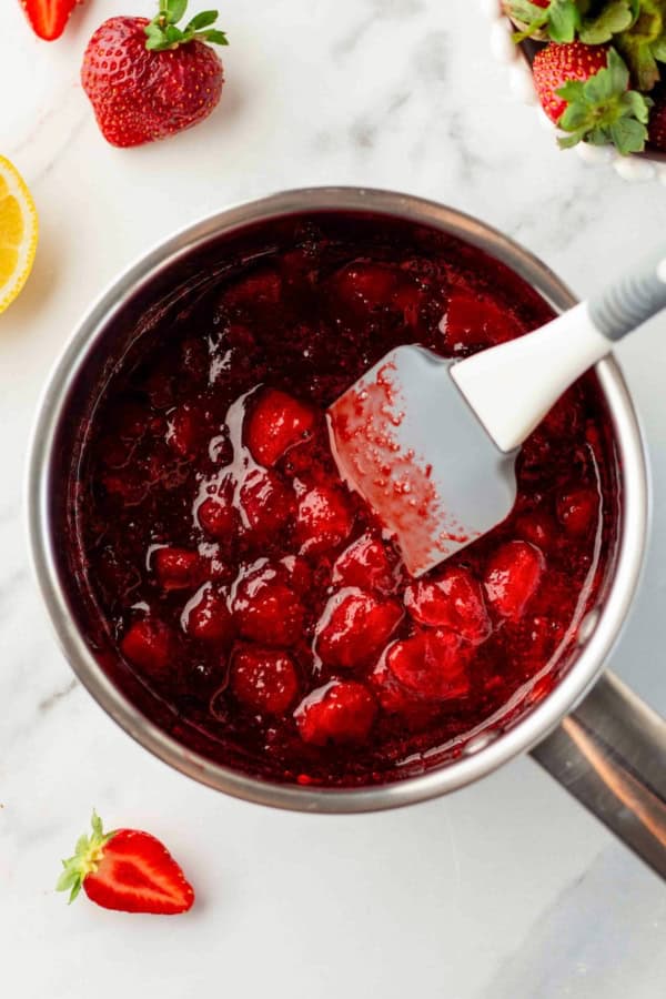 Made with only 4 ingredients in just 10 minutes, this homemade Strawberry Compote recipe is so thick and jammy - with no cornstarch needed! A saucepan filled with chunky strawberry compote being stirred with a spatula on a marble surface.