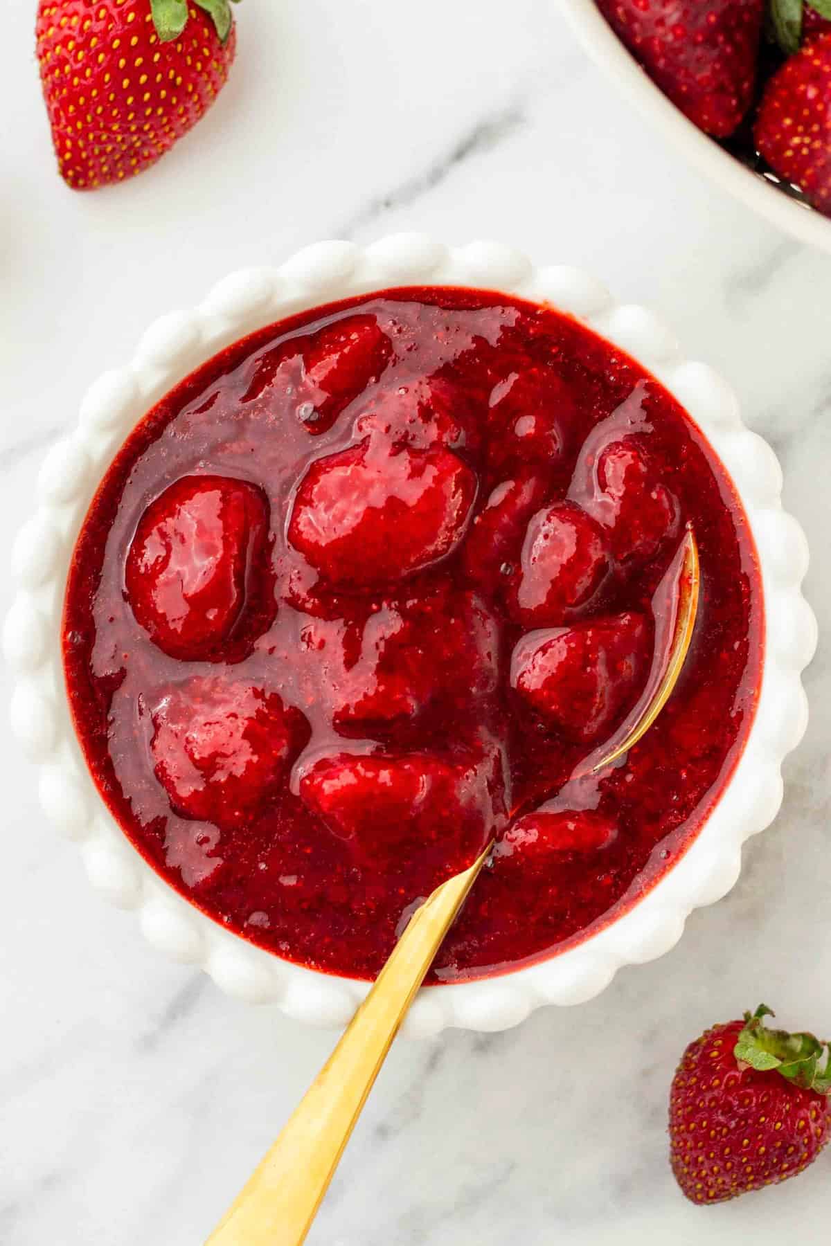 Made with only 4 ingredients in just 10 minutes, this homemade Strawberry Compote recipe is so thick and jammy - with no cornstarch needed! A white bowl filled with thick strawberry compote containing whole strawberries, with a gold spoon inside. Fresh strawberries are placed around the bowl on a marble surface.