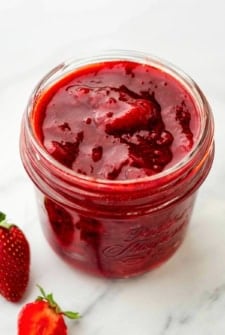 A glass jar filled with thick homemade strawberry compote sits on a white surface, with fresh strawberries placed nearby.