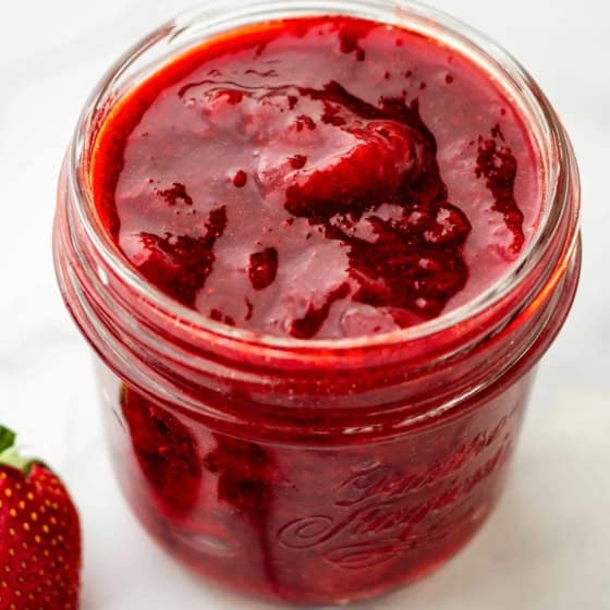 A glass jar filled with thick homemade strawberry compote sits on a white surface, with fresh strawberries placed nearby.