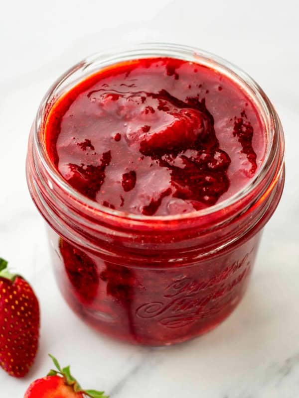 A glass jar filled with thick homemade strawberry compote sits on a white surface, with fresh strawberries placed nearby.