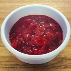 A white bowl filled with thick, glossy strawberry compote sauce, with visible chunks of strawberries, placed on a wooden surface.