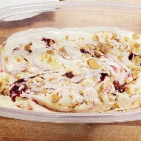 A container of creamy strawberry cheesecake no-churn ice cream swirled with red strawberry sauce and topped with crushed graham crackers sits on a wooden surface.