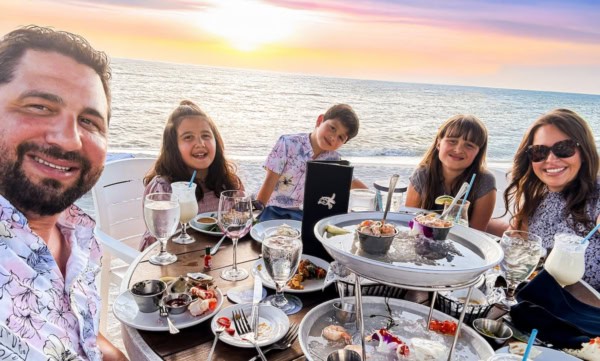 A family of five smiles at a seaside restaurant table during sunset, enjoying seafood and drinks with the ocean in the background.