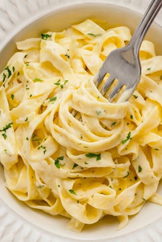 A close-up of creamy homemade Alfredo Sauce on fettuccine pasta garnished with chopped parsley in a white bowl, with a fork twirling some noodles.