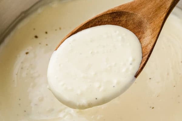 Thick, creamy, homemade Alfredo sauce recipe in 10 minutes with just 5 ingredients and a pinch of nutmeg, the Italian secret most home cooks skip! A wooden spoon holds up a portion of creamy homemade Alfredo sauce, with the rest of the sauce visible in the background. The sauce is smooth with small specks of black pepper.