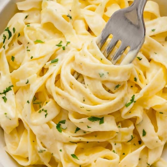 A close-up of creamy homemade Alfredo Sauce on fettuccine pasta garnished with chopped parsley in a white bowl, with a fork twirling some noodles.
