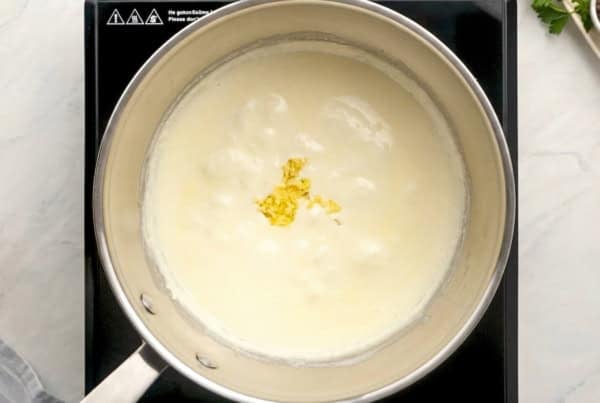 Thick, creamy, homemade Alfredo sauce recipe in 10 minutes with just 5 ingredients and a pinch of nutmeg, the Italian secret most home cooks skip! A saucepan filled with a creamy white pasta sauce is heating on a stovetop, with a small amount of grated garlic in the center of the sauce.