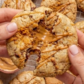 Close-up of hands pulling apart a gooey Biscoff stuffed cookie, revealing a melted cookie butter filling. Several cookies with Biscoff drizzle rest on a cooling rack in the background.