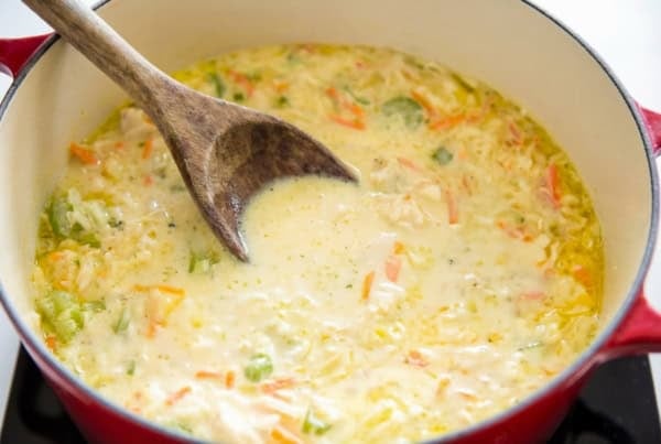 The best homemade Chicken and Dumplings recipe you'll ever make! Ready in under an hour with a thick creamy broth, juicy chicken, and big fluffy dumplings. A wooden spoon rests in a pot of creamy soup with visible pieces of carrots, celery, and onion, all simmering together in a red Dutch oven on a stovetop.