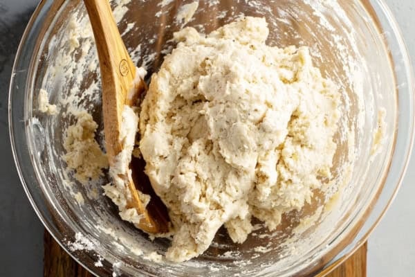 The best homemade Chicken and Dumplings recipe you'll ever make! Ready in under an hour with a thick creamy broth, juicy chicken, and big fluffy dumplings. A glass mixing bowl with thick, lumpy dumpling dough being stirred by a wooden spoon, set on a wooden board.