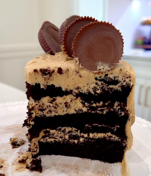 A slice of layered chocolate cake with creamy frosting, topped with two large chocolate peanut butter cups, sits on a white plate with cake crumbs around it.