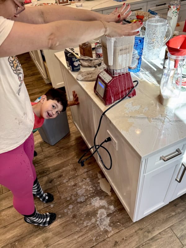 A woman tries to stop a blender overflowing with liquid, spilling onto the kitchen counter and floor. A smiling child peeks from behind the island, enjoying the messy scene. The kitchen is cluttered with ingredients and utensils.
