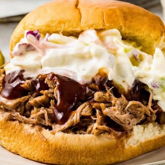 An instant pot pulled pork sandwich topped with barbecue sauce and creamy coleslaw on a bun, served on a white plate with a pickle spear on the side.