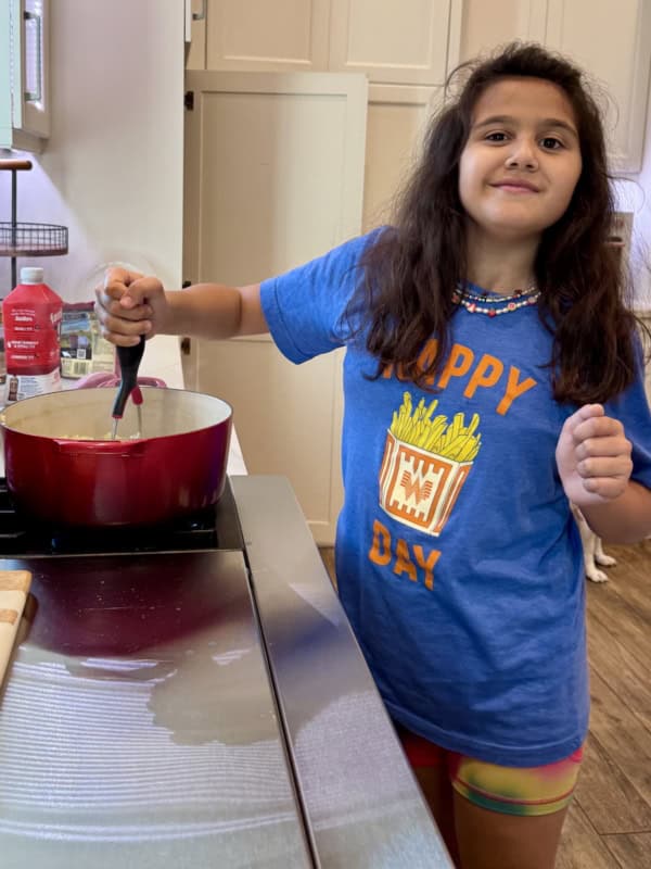 A young girl with long dark hair stirs food in a red pot on a stove. She wears a blue Happy Day t-shirt with a fries graphic and colorful shorts, standing in a bright kitchen and smiling at the camera.