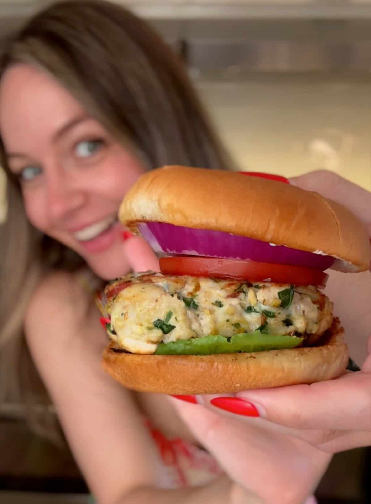 A woman with long hair smiles while holding a chicken burger with red onion, lettuce, and a golden bun close to the camera, showing off the sandwich in focus.
