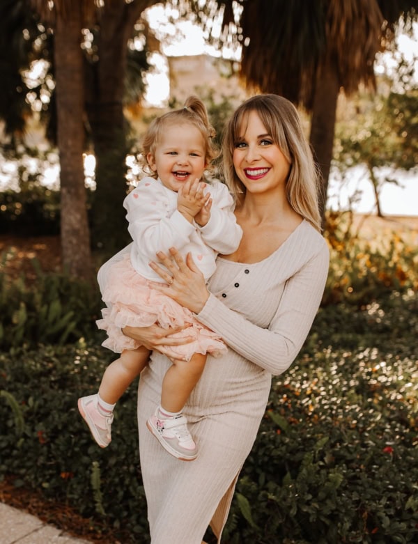 A smiling woman in a light-colored dress holds a happy toddler wearing a white sweater and pink skirt. They stand outdoors in a garden with trees and greenery in the background.