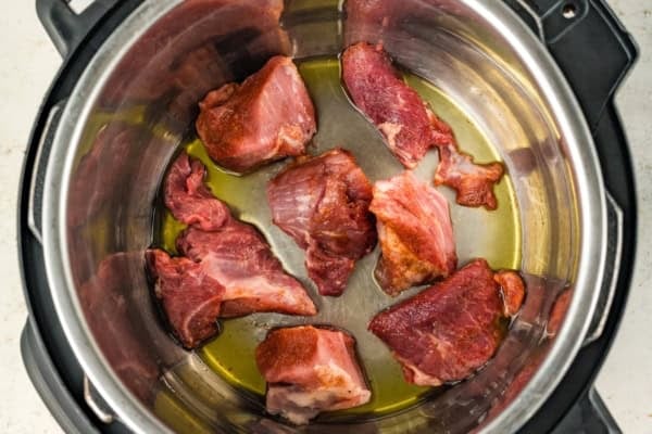 Fall-apart tender Instant Pot pulled pork recipe with a smoky brown sugar rub and tangy BBQ sauce. Done in 90 minutes, no smoker required! Chunks of raw pork are searing in oil at the bottom of a stainless steel Instant Pot pressure cooker. The meat pieces are spaced out and beginning to brown.
