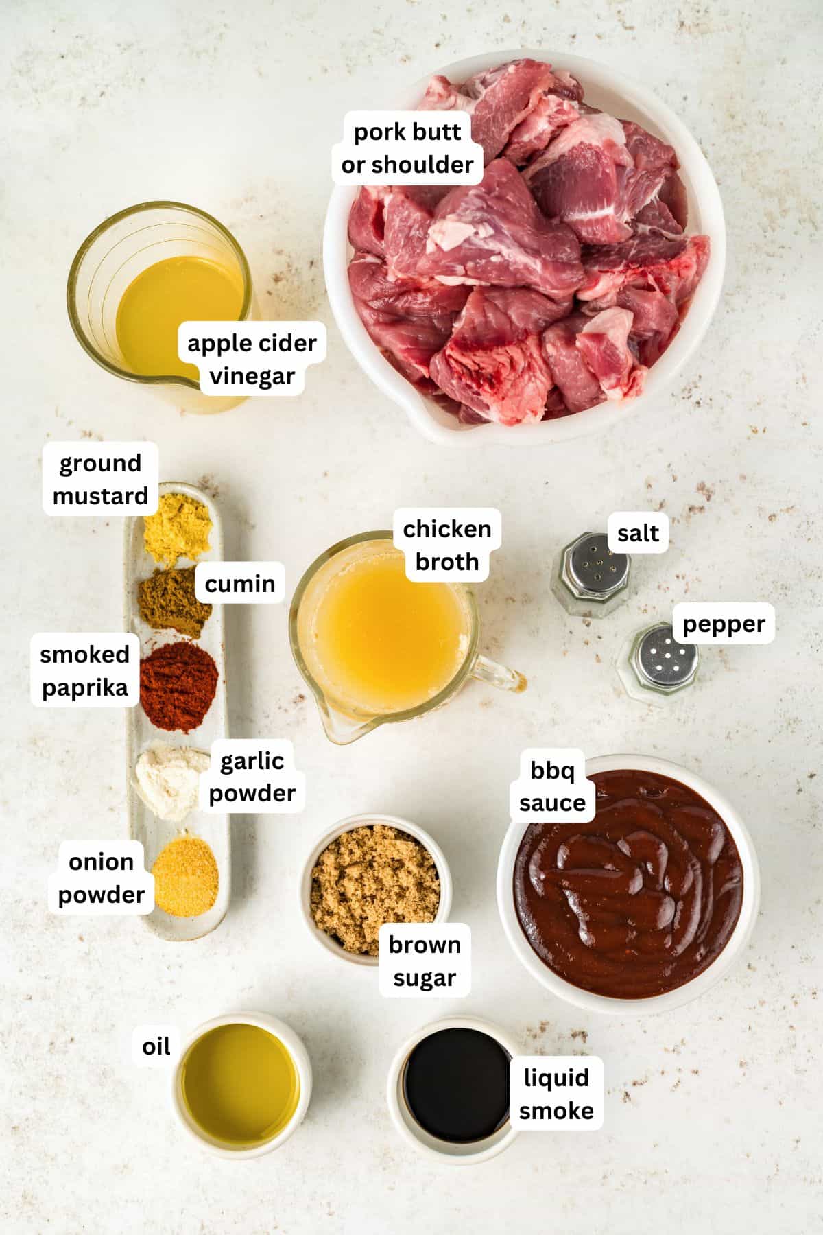 Fall-apart tender Instant Pot pulled pork recipe with a smoky brown sugar rub and tangy BBQ sauce. Done in 90 minutes, no smoker required! Labeled ingredients for pulled pork instant pot recipe are arranged on a white surface, including pork butt, chicken broth, apple cider vinegar, bbq sauce, brown sugar, spices, oil, liquid smoke, salt, and pepper.