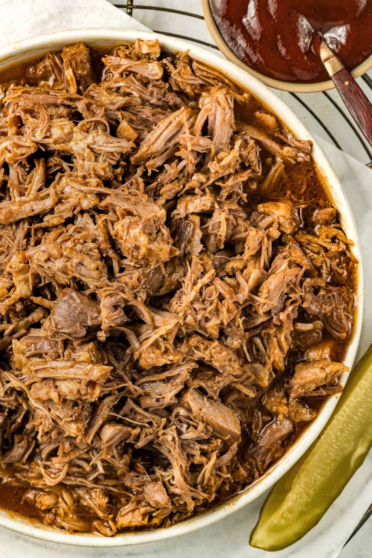 Fall-apart tender Instant Pot pulled pork recipe with a smoky brown sugar rub and tangy BBQ sauce. Done in 90 minutes, no smoker required! A large serving bowl filled with shredded instant pot pulled pork in bbq sauce, with a pickle spear and a small bowl of extra barbecue sauce on the side.