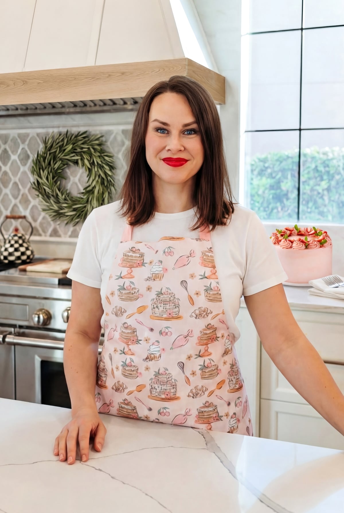 A woman with brown hair and red lipstick stands in a modern kitchen, wearing a white shirt and a pink apron with cake and baking designs. She smiles at the camera, and a decorated cake is visible on the counter behind her.