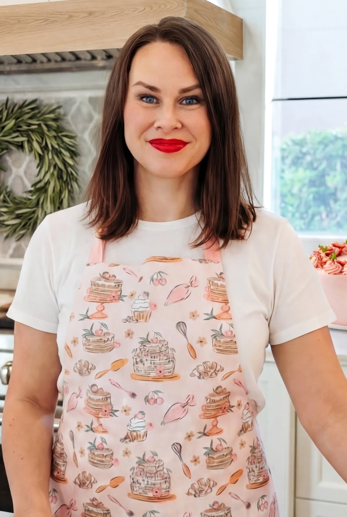 Jessica Segarra smiles at the camera. She is wearing a white shirt and a pink apron decorated with cake and baking illustrations. The background shows a kitchen setting.