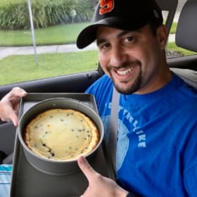 A smiling man wearing a black cap and blue t-shirt sits in a car and holds a round cheesecake with chocolate chips in a baking pan on his lap.