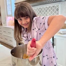 A young girl in pajamas stirs cookie dough in a metal mixing bowl on a kitchen counter, smiling at the camera. She stands in a bright, modern kitchen with white cabinets and a patterned backsplash.