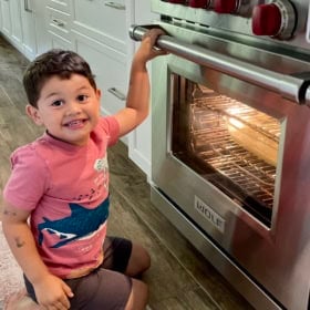 A young boy kneels on a kitchen floor, smiling at the camera while holding the handle of a stainless steel oven with a loaf of bread baking inside. The ovens interior is lit, and the kitchen has white cabinets.