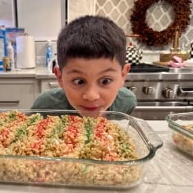 A young boy with wide eyes excitedly looks at a glass baking dish filled with Rice Krispies treats decorated with red and green sprinkles, on a kitchen counter with a stove and holiday wreath in the background.
