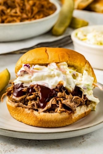An instant pot pulled pork sandwich topped with barbecue sauce and creamy coleslaw on a plate, with a pickle spear and a bowl of coleslaw in the background.
