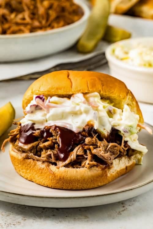 An instant pot pulled pork sandwich topped with barbecue sauce and creamy coleslaw on a plate, with a pickle spear and a bowl of coleslaw in the background.