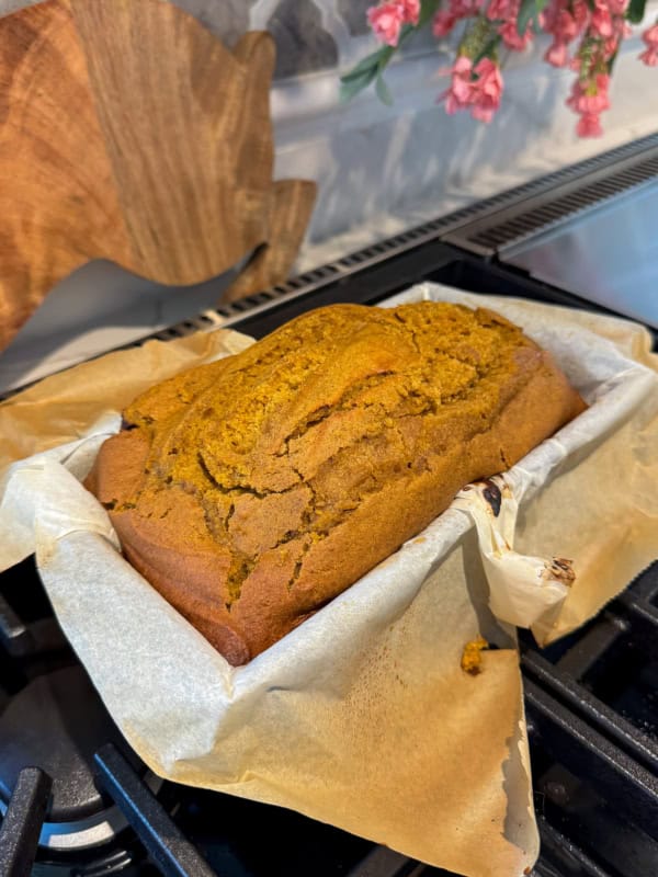 A freshly baked loaf of bread with a golden-brown crust sits in a parchment-lined loaf pan on a stove, with wooden and floral decor visible in the background.