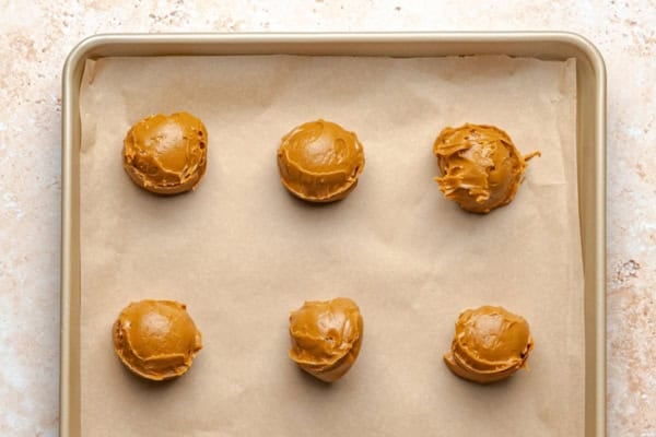 Biscoff Stuffed Cookie Butter Cookies with a molten cookie butter center, crushed Biscoff cookies, and dark chocolate are for serious Biscoff lovers! Six scoops of biscoff cookie butter are arranged in two rows on a parchment-lined baking sheet, ready to be frozen.