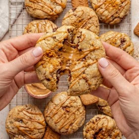 Hands breaking a large stuffed biscoff cookie butter cookie drizzled with biscoff cookie butter over a cooling rack filled with similar cookies and Biscoff biscuits.