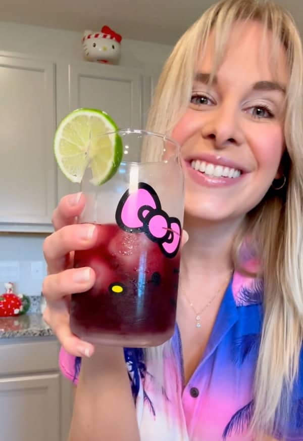 A smiling woman in a pink and blue shirt holds up a Hello Kitty glass with a dark red drink, ice, and a lime slice. She stands in a kitchen with light cabinets and Hello Kitty decorations in the background.