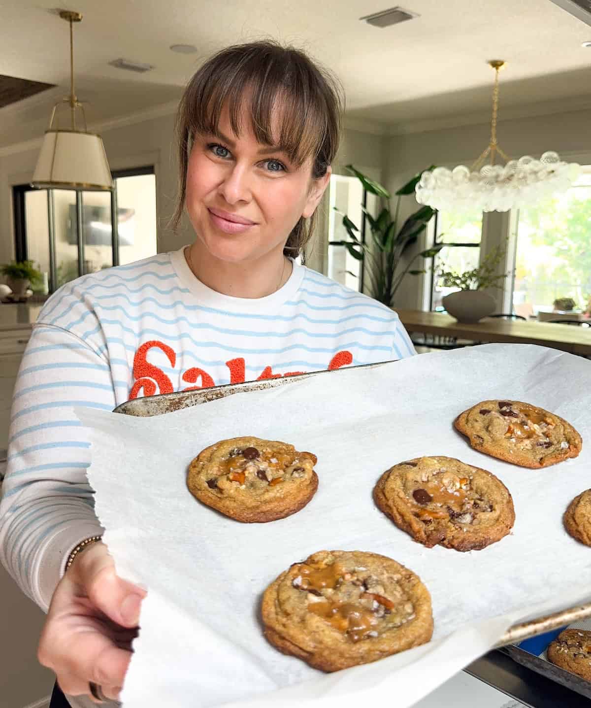 The ultimate sweet and salty kitchen sink cookies with a brown butter base, gooey caramel, double chocolate, pretzels, chips, and a flaky sea salt finish. A woman in a striped shirt smiles while holding a tray of freshly baked Everything But The Kitchen Sink Cookies on parchment paper in a bright, modern kitchen.