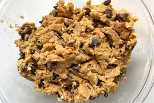 The ultimate sweet and salty kitchen sink cookies with a brown butter base, gooey caramel, double chocolate, pretzels, chips, and a flaky sea salt finish. A close-up of raw kitchen sink cookie dough with visible chocolate chips, potato chip and pretzel pieces.